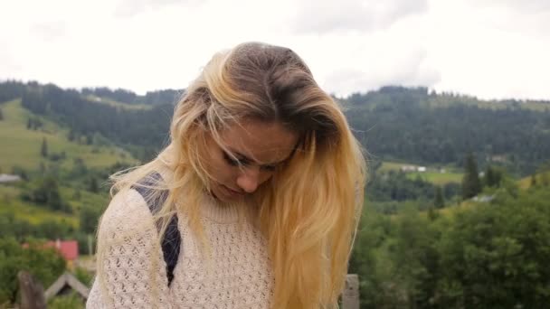 Hermosa chica posando ante la cámara y sonriendo sobre un fondo de bosque y pueblo de montaña — Vídeo de stock