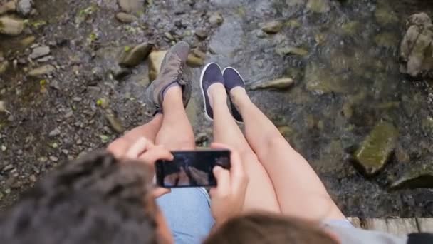 Fiatal pár turisták hátizsák egy hídon, egy hegyi folyó. Csinál egy fénykép, a lábak, a háttérben folyó — Stock videók