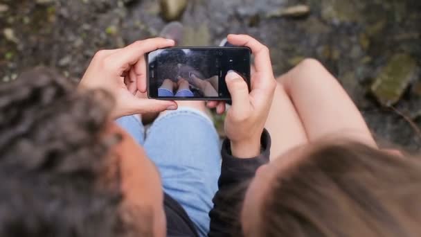 Pareja joven de turistas con mochilas en un puente sobre un río de montaña. Hacer una foto de los pies en el fondo del río — Vídeo de stock