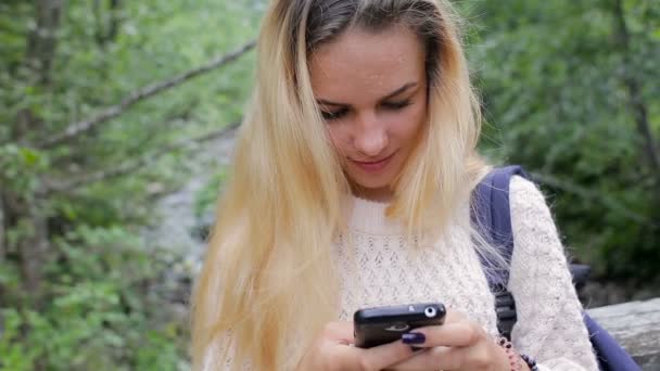 Ung flicka med en telefon på en bro över en flod i berg — Stockvideo