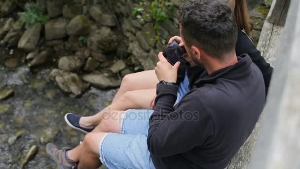 Giovane coppia di turisti con zaini su un ponte su un fiume di montagna. Fai una foto dei piedi sullo sfondo del fiume — Video Stock