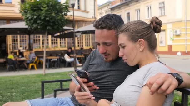 Feliz pareja joven sentada en un banco y usando un teléfono inteligente. Mujer y hombre viendo vídeo, fotos — Vídeos de Stock