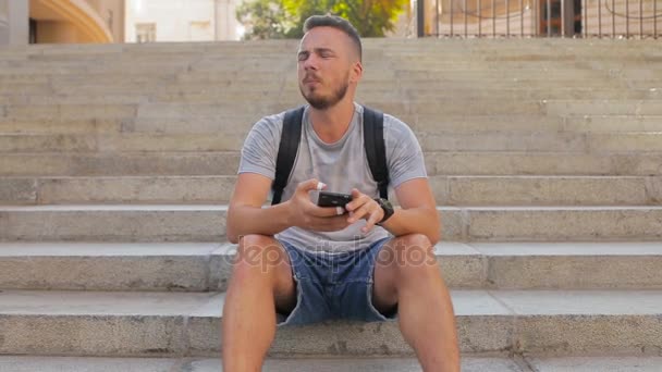 Young guy sits on the steps in the city and plays a game on the phone — Stock Video