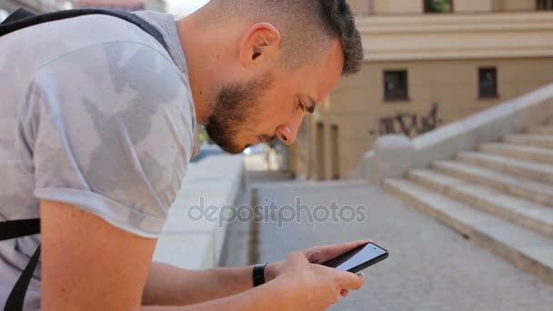 Jonge kerel zit op de stappen in de stad en een spel speelt op de telefoon — Stockvideo