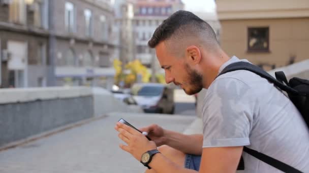 Jovem senta-se nos degraus da cidade e joga um jogo no telefone — Vídeo de Stock