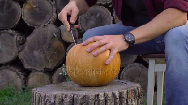 Man kerft van een pompoen Jack-o-lantaarn in de achtertuin op een boomstronk — Stockvideo