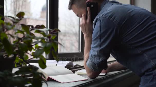 Serpenyőben a fiatal férfi kap segítséget a tanulás telefon állva az ablak mellett — Stock videók
