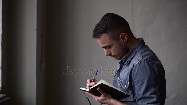 Medium shot of the guy writes in the notebook while standing near the window opposite the gray wall — Stock Video