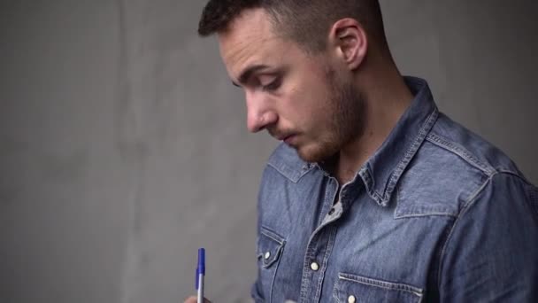 PAN del tipo escribe en el cuaderno mientras está de pie cerca de la ventana frente a la pared gris — Vídeos de Stock