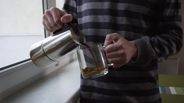 Primer plano de un joven barbudo vertiendo café en una taza de pie cerca de la ventana en la cocina — Vídeos de Stock