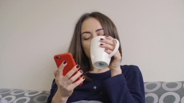 Joven mujer sonriente bebiendo café en casa en su cama y comprobando el teléfono inteligente — Vídeos de Stock