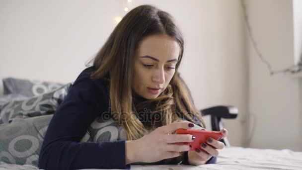 Vrolijke jonge meisje liggen onder deken en typen op de mobiele telefoon in bed — Stockvideo