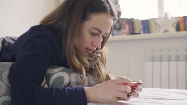 自宅のベッドを横になっているスマート フォンでゲームを演奏若い女性 — ストック動画