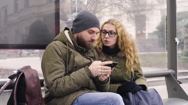 Joven hombre y mujer usar el teléfono móvil mientras espera en una parada de autobús — Vídeo de stock