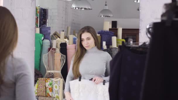 Una joven elige un paño para un vestido cerca de un espejo en el taller — Vídeos de Stock