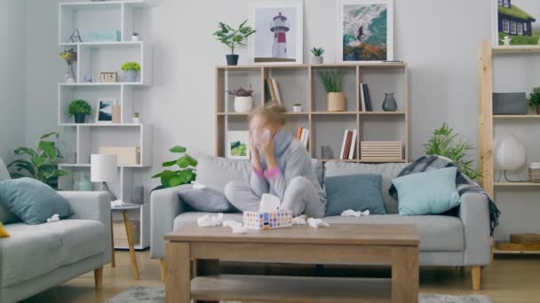 Girl with a cold sneezes while sitting on a couch in a room — Stock Video