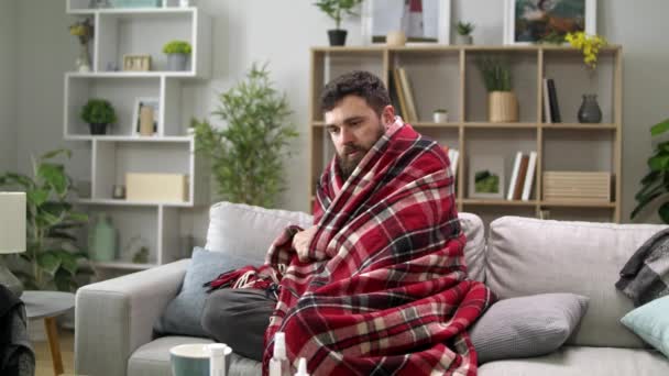 Nemocný muž s zimnicí a zabalí se do deky na pohovce v obýváku — Stock video