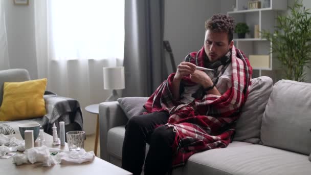 Ένας άρρωστος άνθρωπος μετράει τη θερμοκρασία στο σπίτι στον καναπέ. — Αρχείο Βίντεο