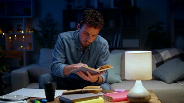 Genç adam akşam evde çalışırken bir kitap okur. — Stok video