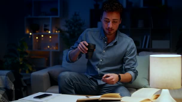 Un joven bebe café y sigue estudiando en casa por la noche. . — Vídeos de Stock