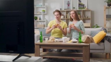 Genç bir çift hamburger yiyor ve oturma odasında televizyon izliyor.