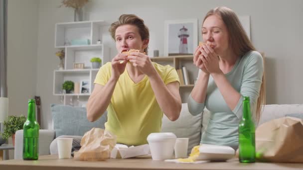 Mladý pár jíst hamburger a dívat se na televizi v obývacím pokoji — Stock video