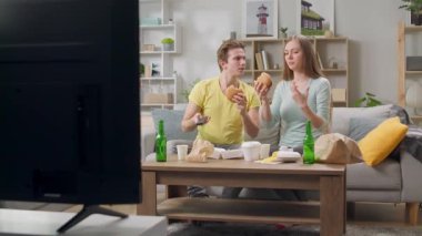 Genç bir çift hamburger yiyor ve oturma odasında televizyon izliyor.