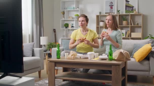 Jeune couple manger un hamburger et regarder la télévision dans le salon — Video