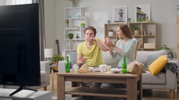 Mladý pár jíst hamburger a dívat se na televizi v obývacím pokoji — Stock video