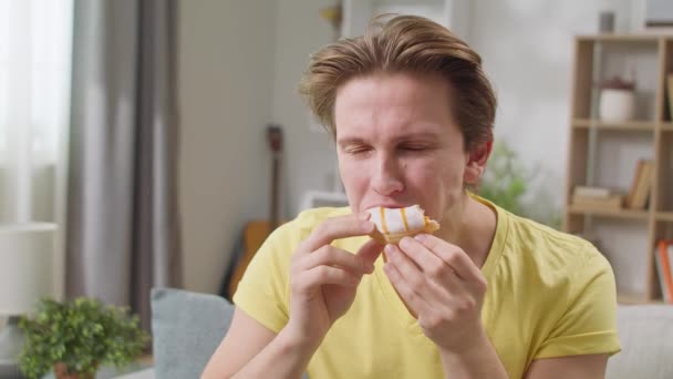 Cheerful guy appetizingly eats a donut at home on the sofa — Stock Video