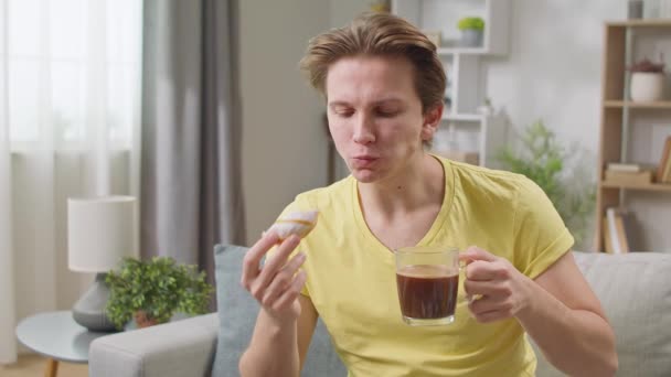 Chico alegre come un donut en casa en la sala de estar y bebe café — Vídeos de Stock
