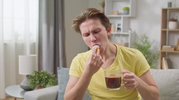 Веселий хлопець їсть пончик вдома у вітальні і п'є каву — стокове відео