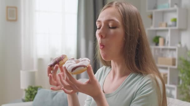 Веселая девушка аппетитно ест дома два пончика на диване — стоковое видео