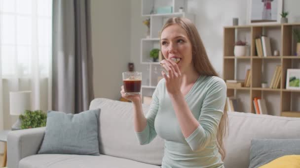 Vrolijke vrouw eet een donut thuis in de woonkamer en drinkt koffie — Stockvideo