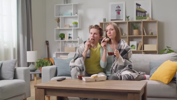 Νεαρό ζευγάρι βλέπει τηλεόραση και πίνει καφέ με ντόνατς κρυμμένοι σε μια κουβέρτα — Αρχείο Βίντεο