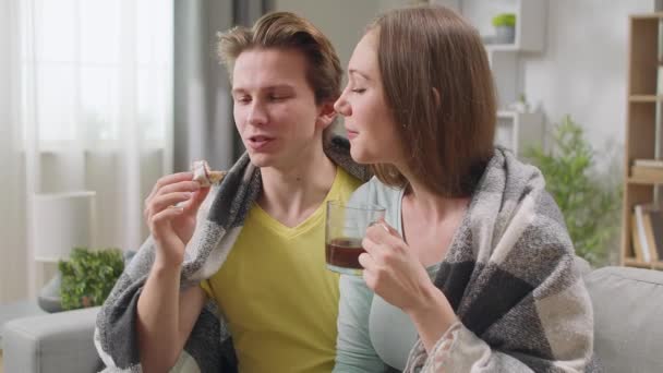 Jeune couple regardant la télévision et buvant du café avec des beignets cachés dans une couverture — Video