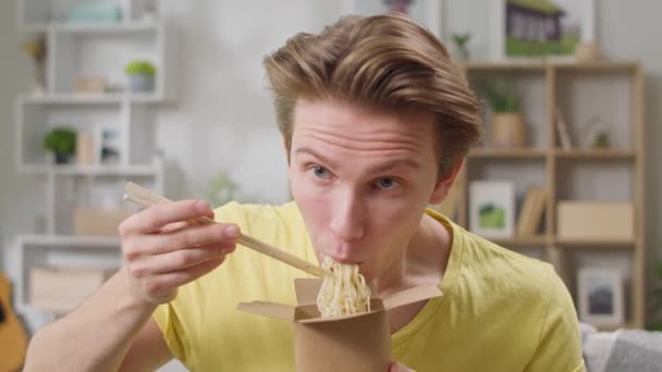 Joven comiendo fideos de caja con palillos en casa en la sala de estar — Vídeos de Stock