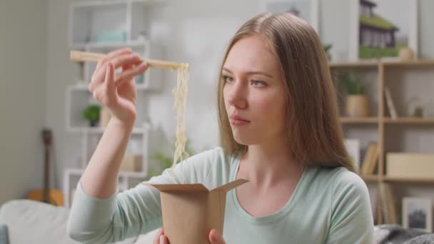 Mujer joven comiendo fideos de una caja con palillos en casa en un sofá — Vídeos de Stock