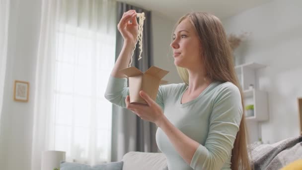 Γυναίκα τρώει νουντλς από ένα κουτί με ξυλάκια στο σαλόνι. — Αρχείο Βίντεο