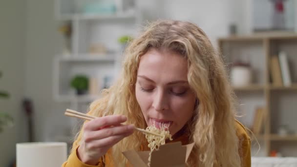 Mujer joven comiendo fideos de una caja con palillos en casa en un sofá — Vídeos de Stock