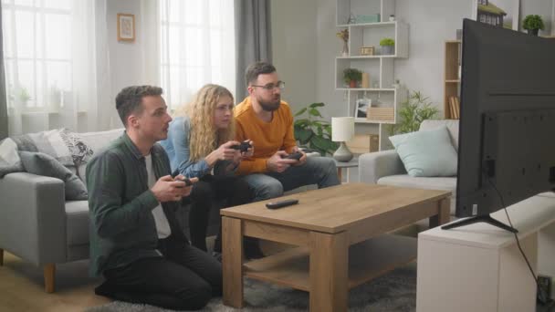 Трое друзей играют в видеоигры, сидя на диване в гостиной — стоковое видео