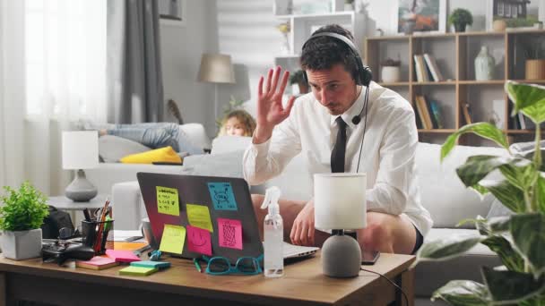 A young man begins a video conference. And his girlfriend is resting on a sofa in the background. Work at home. — Stock Video