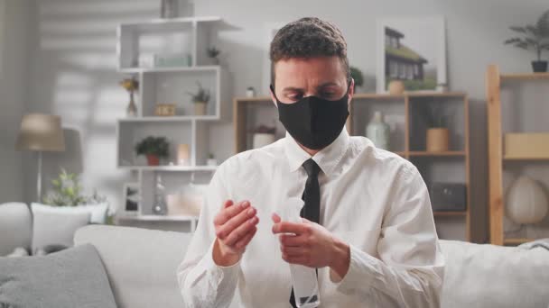 Jonge zakenman zit thuis op een bank in een medisch masker behandelt handen met een ontsmettingsmiddel — Stockvideo