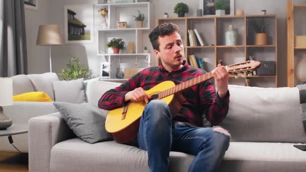 Młody człowiek gra na gitarze w domu na kanapie. — Wideo stockowe