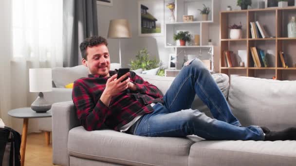 Mladý muž používá chytrý telefon doma, zatímco leží na gauči — Stock video