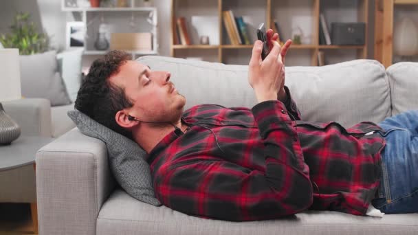 A young man listens to music and dances in headphones while lying on a sofa — Stock Video