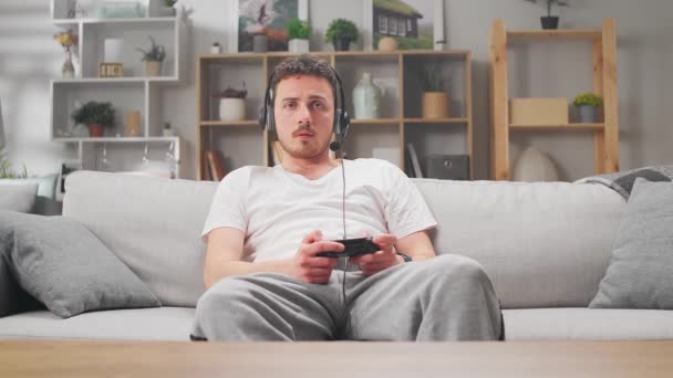 Een jonge man speelt online video games met een koptelefoon op de bank — Stockvideo