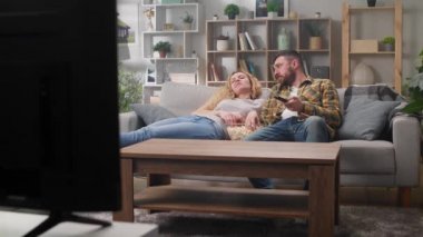 Oturma odasında kanepede uzanmış televizyon seyreden ve patlamış mısır yiyen genç bir çift.