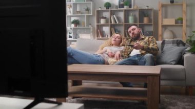 Oturma odasında kanepede uzanmış televizyon seyreden ve patlamış mısır yiyen genç bir çift.