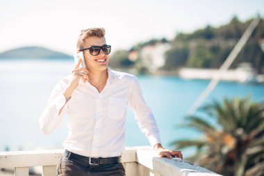 Denizin panoramik manzaralı lüks resort Hotel'de konaklamanın tadını yakışıklı delikanlı. Kazanılan bir tropikal tatil onun smartphone seçenekleri kullanarak gülümseyen iş adamı. İş seyahat. Dış kaynak